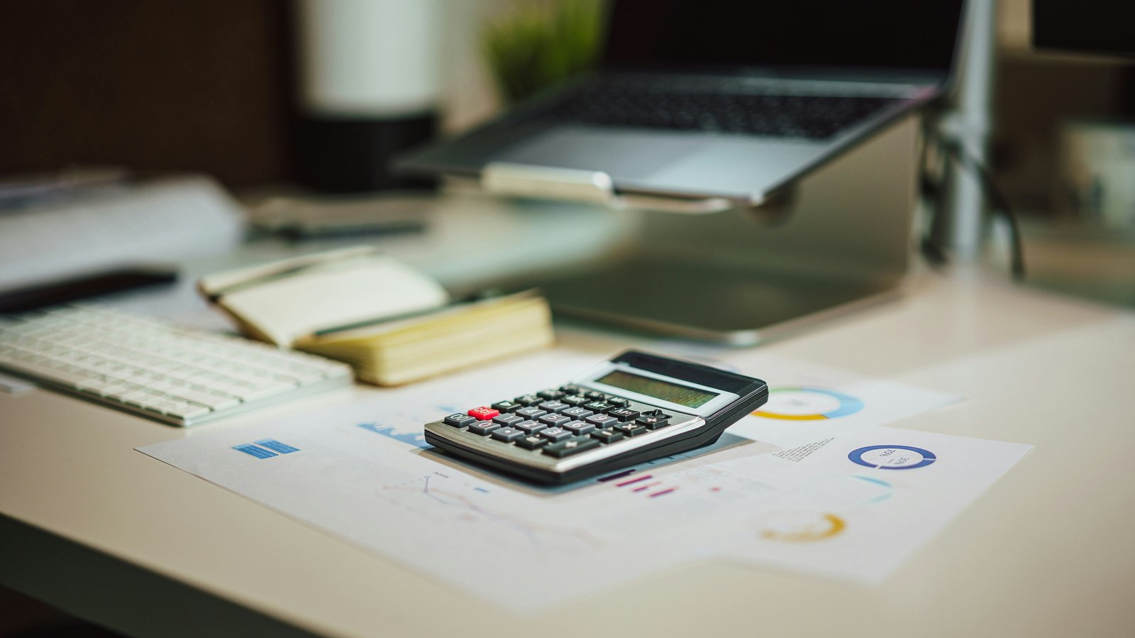 A calculator resting on financial charts next to a laptop on a desk